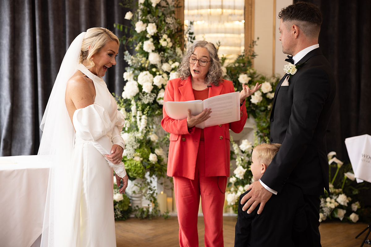 Bride and groom sharing a laugh during their wedding ceremony with a floral fireplace backdrop