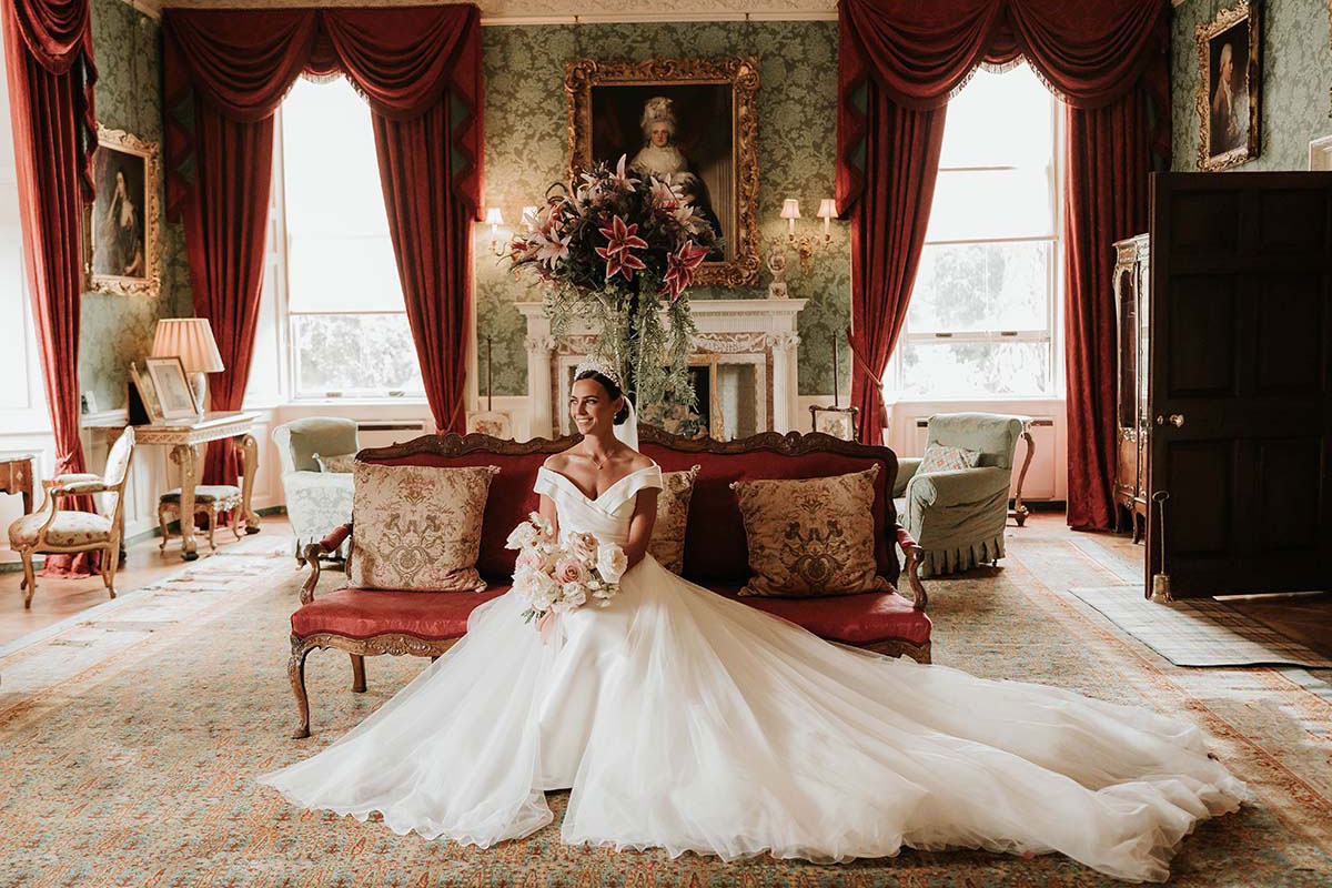 Bride in an off-the-shoulder gown sitting on a red sofa in an ornate room with red curtains and antique furnishings