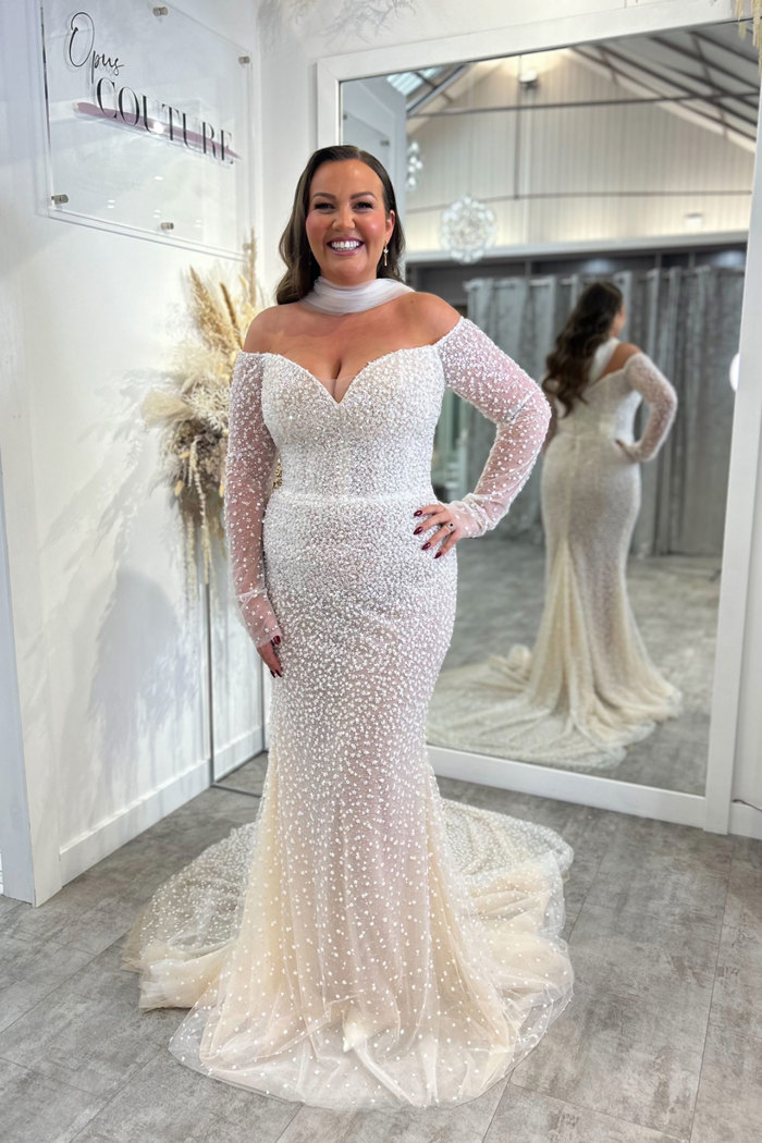 smiling dark-haired model wears off the shoulder long sleeve shimmery wedding dress and a tulle neck scarf