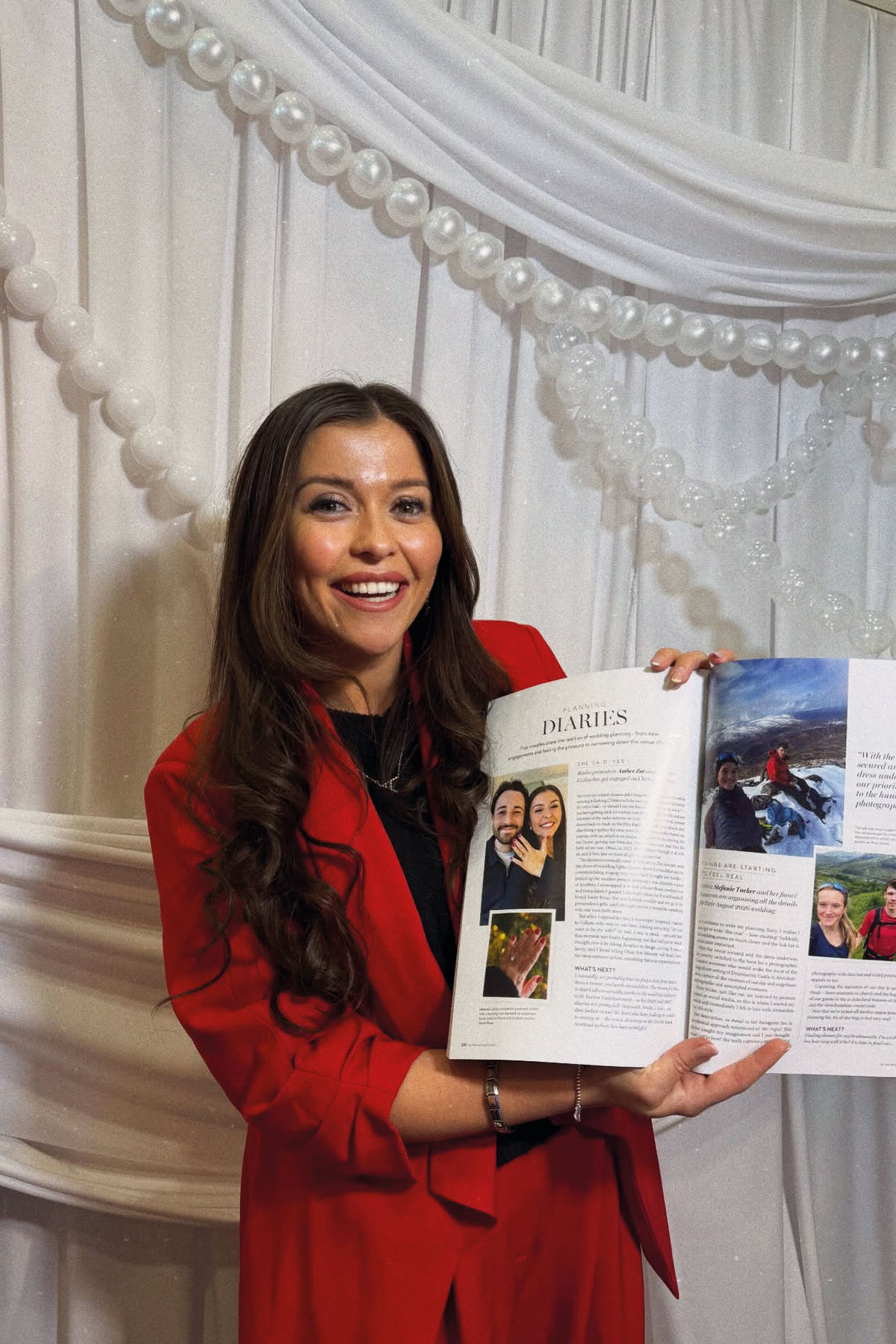 woman in red blazer holding open wedding magazine at scottish wedding show stand with white draped backdrop and balloon decor
