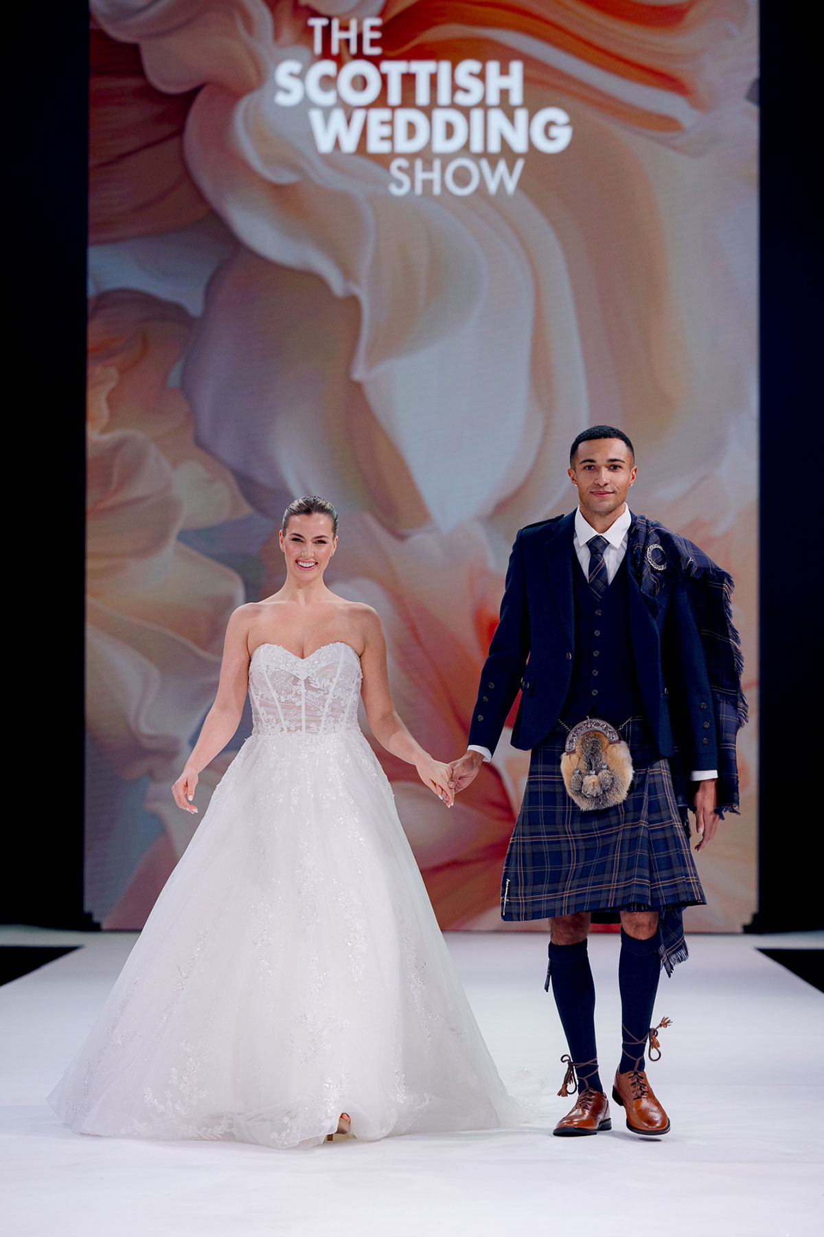 two models on the catwalk at The Scottish Wedding Show, one wearing a strapless wedding dress and the other wearing a kilt outfit