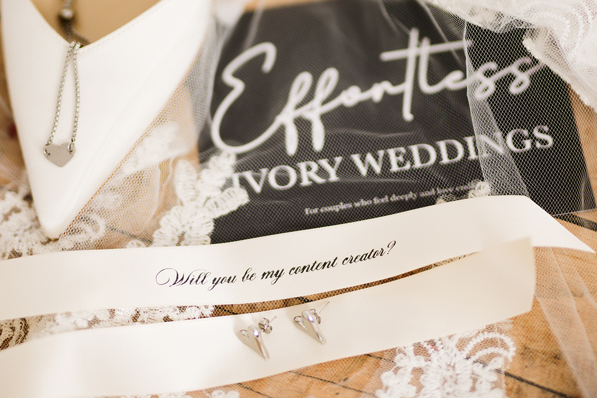 Wedding detail flat lay with bridal shoes, lace veil, silver jewellery and a “Will you be my content creator?” card for Effortless Ivory Weddings.