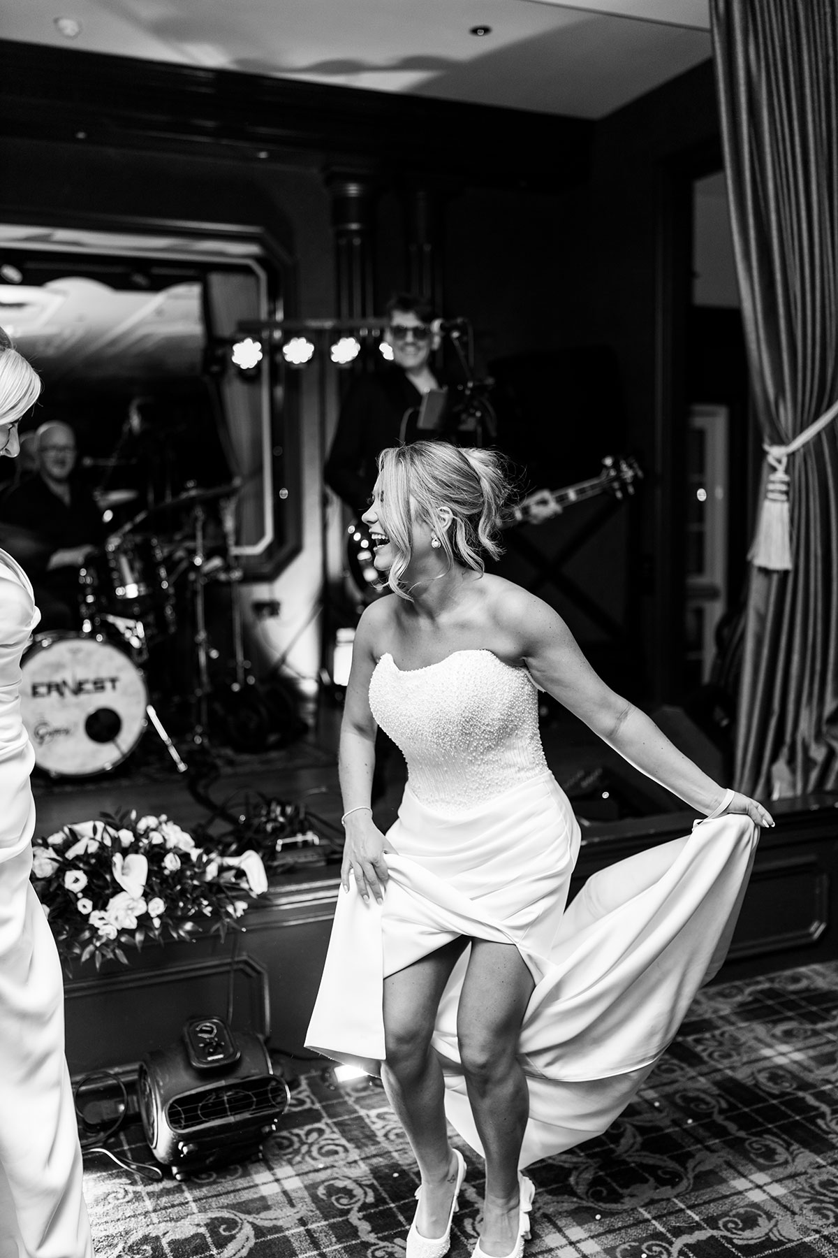 Black and white photograph of one bride dancing and lifting the skirt of her gown while the live band performs behind her.