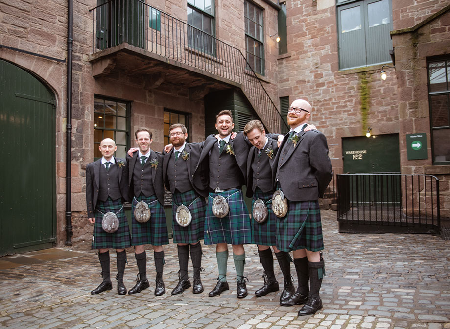 six men in kilts standing on a cobbled pavement surrounded by brown brick buildings