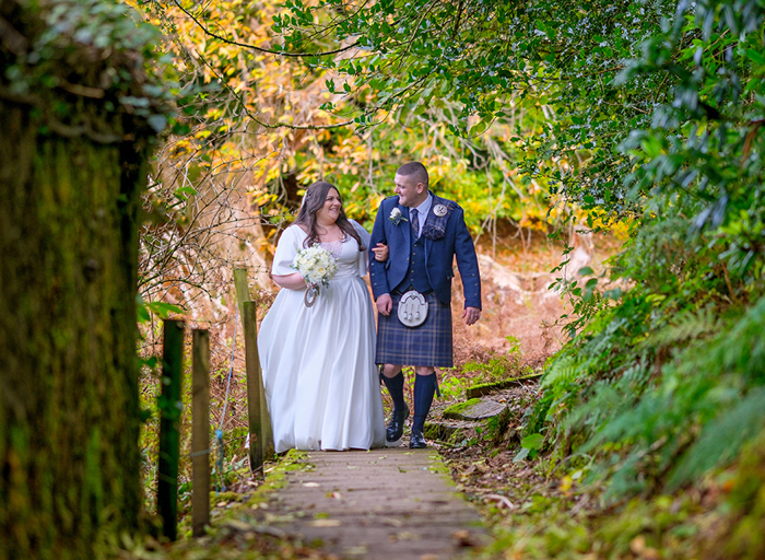 a bride and groom walk along a path holding hands in a woodland area