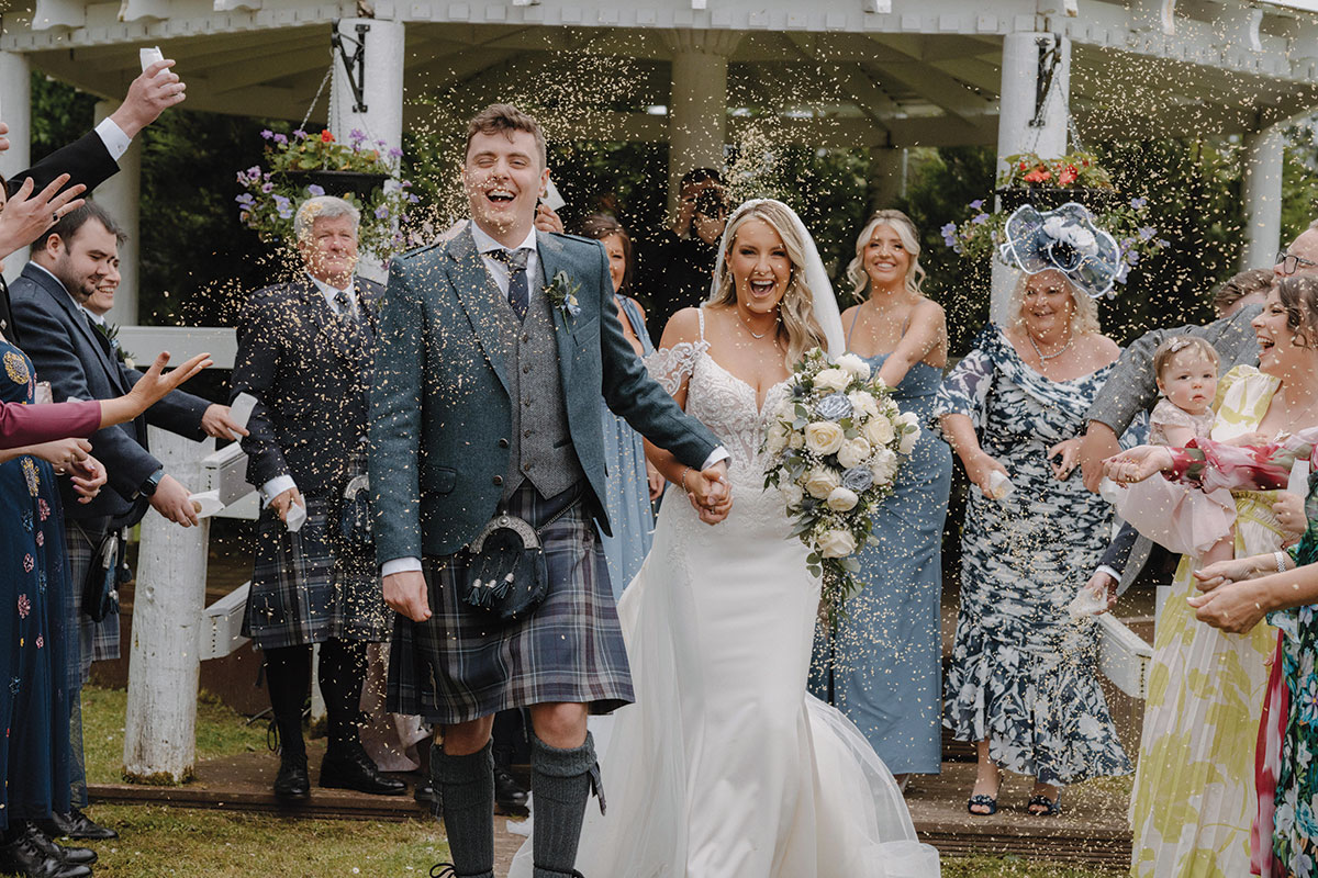 guests through confetti over bride and groom who are smiling and holding hands outside cornhill castle in biggar