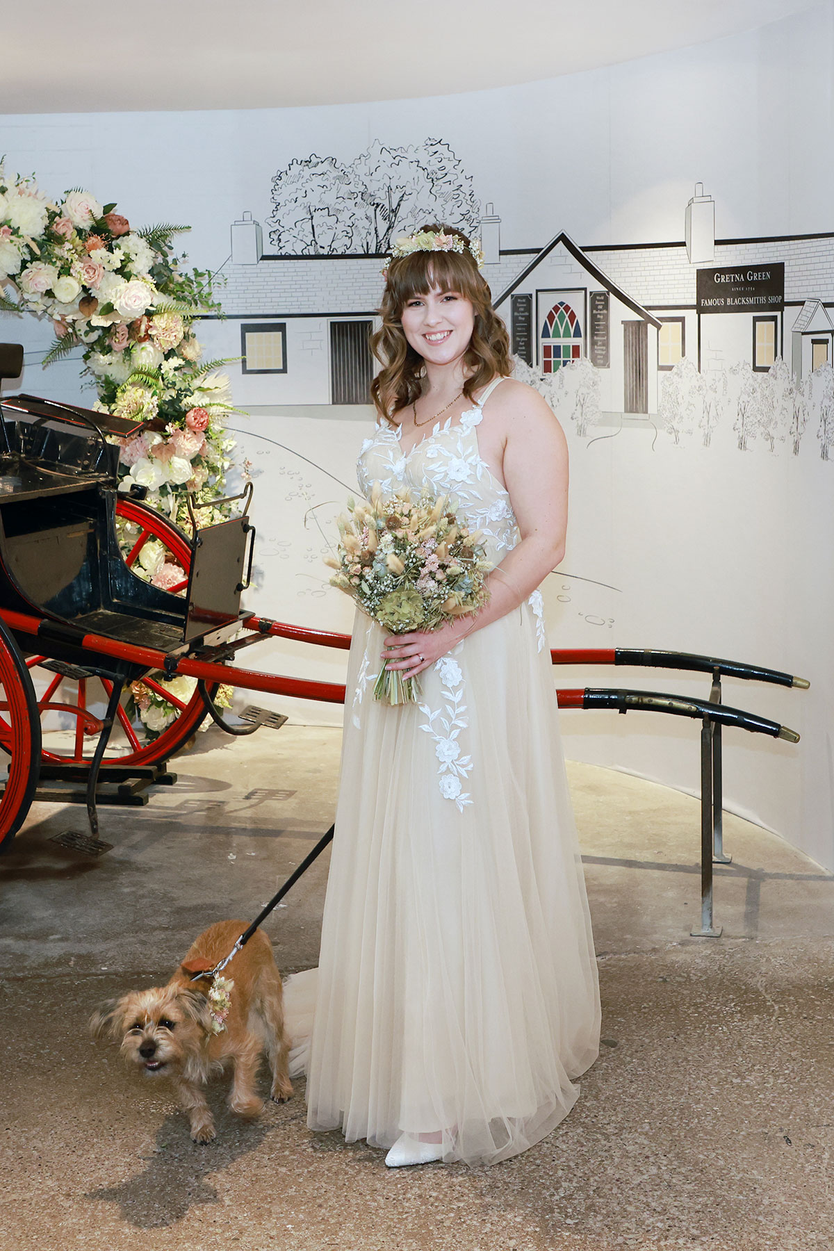 bride wearing a tulle and embroidered wedding dress smiles while holding dog on a lead