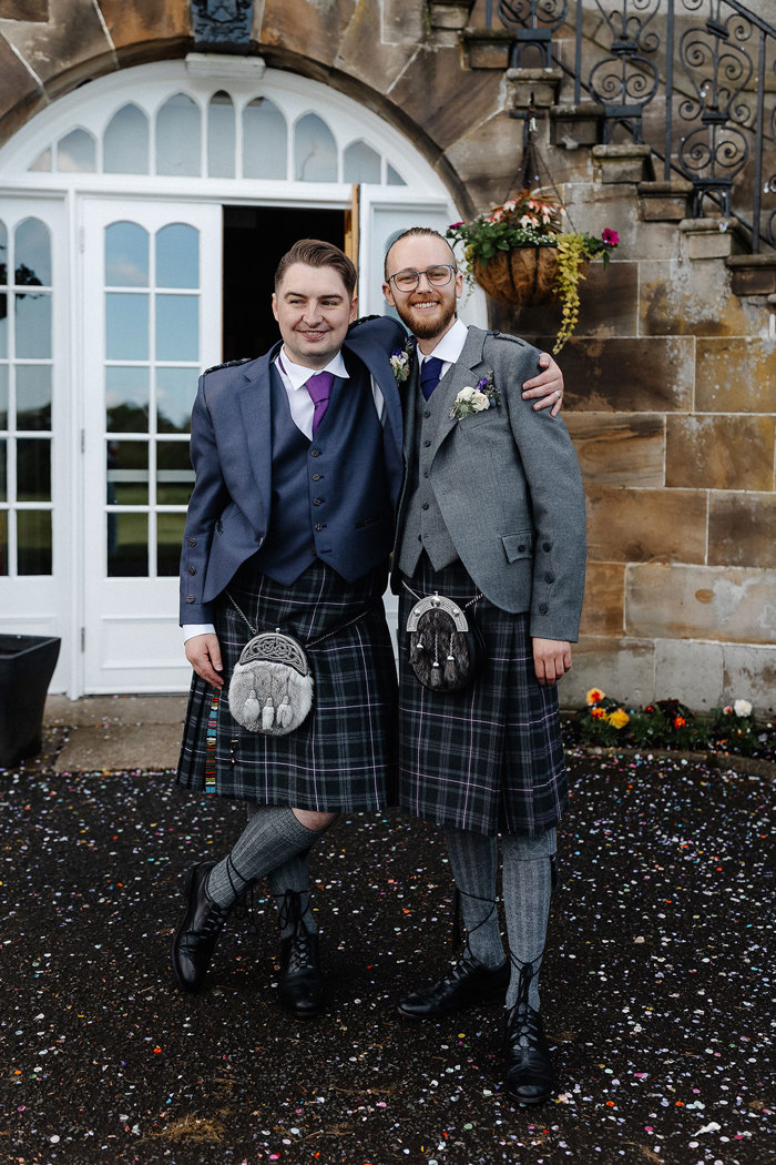 groom and groomsman both in full kilt outfits stand side by side with arms around one another and loads of colourful confetti on the ground