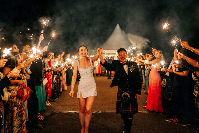 bride and groom smile and hold hands as guests hold sparklers during wedding at mar hall