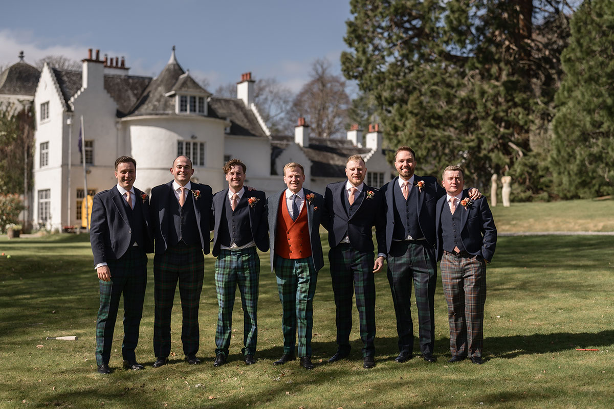 group of groomsmen and groom smile for a photograph all wearing tartan trews