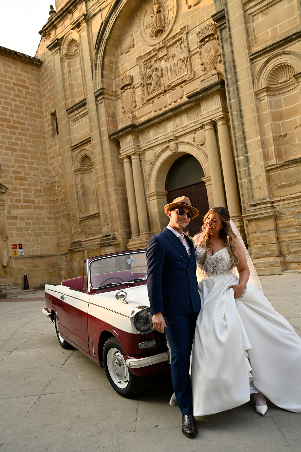 groom in blue suit and brown fedora and sunglasses and bride in ballgown wedding dress with trailing veil stand in front of stationary red and white convertible car with a large intricate brick building behind them
