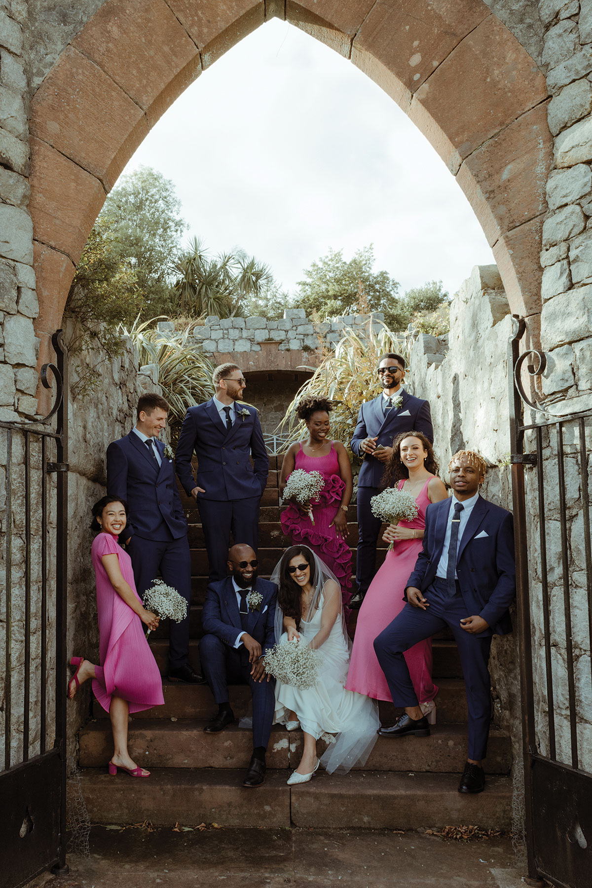 bride and groom sit on steps at walled garden at dougarie boathouse with bridesmaids wearing pink and groomsmen standing next to them