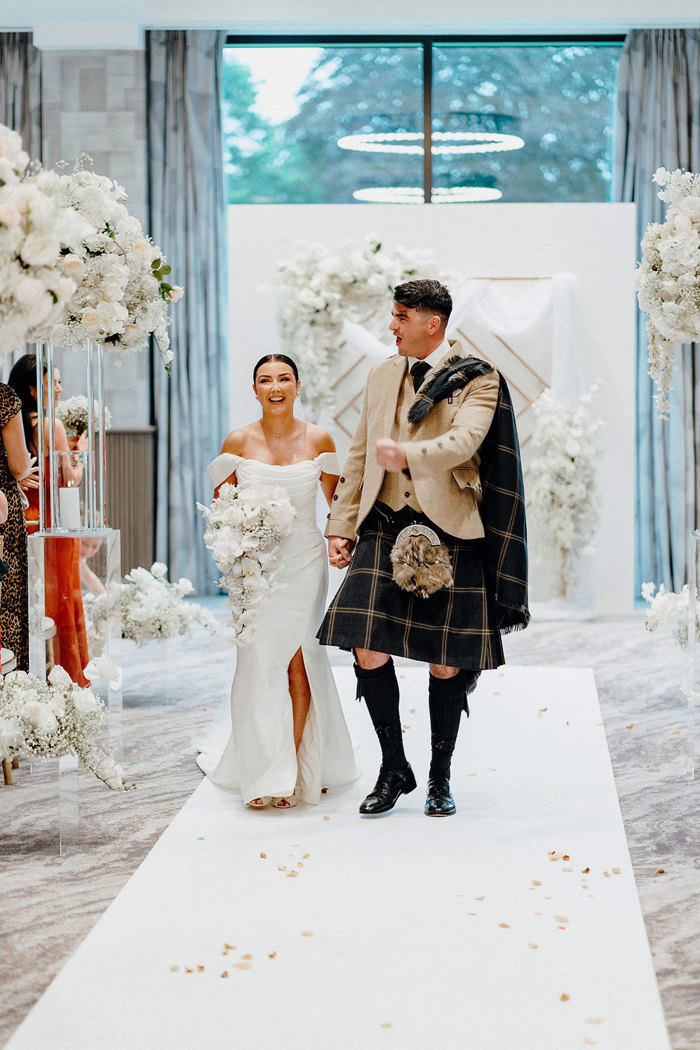 bride in strapless wedding dress with off the shoulder straps holds trailing bouquet while walking up white carpeted aisle with tall groom in beige and dark green kilt outfit who excitedly fist bumps the air