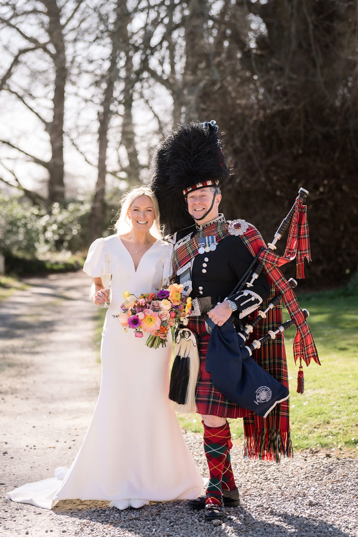 bride wearing a fitted wedding dress with puff sleeves stands next to piper at achnagairn castle