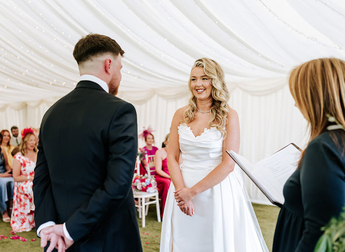 a nervously smiling bride looks at a groom during a wedding ceremony. Guests sitting in rows on chiavari chairs look on and an officiant stands in front of the couple