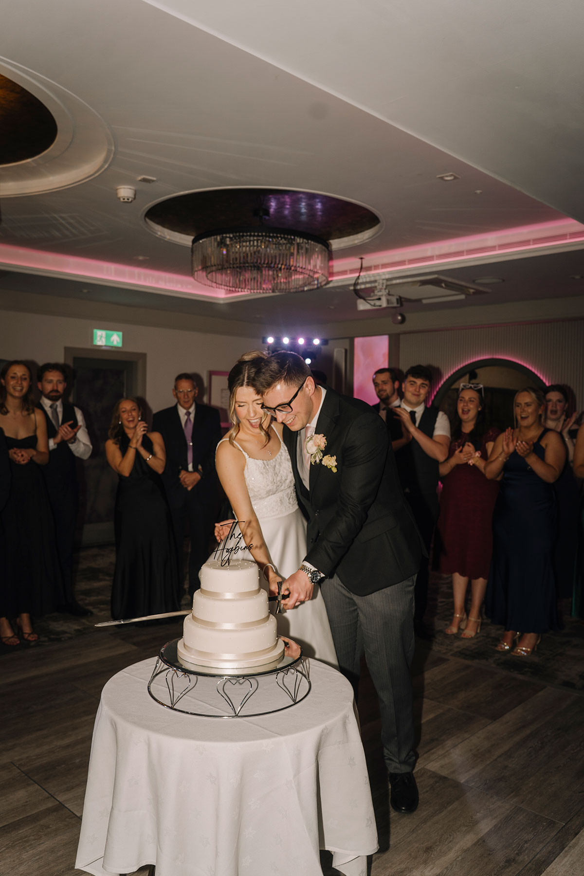 Bride and groom cutting their tiered wedding cake while guests watch and celebrate during the evening reception