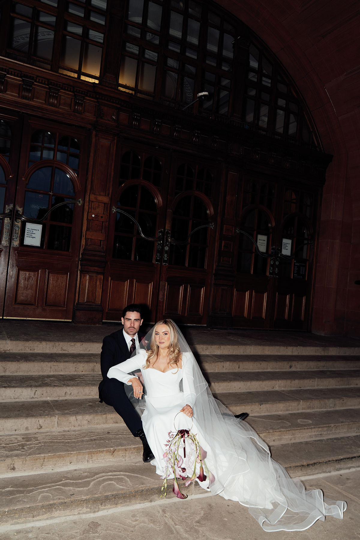 Bride and groom posing on stone steps with the bride holding a sculptural floral hoop bouquet of trailing calla lilies.