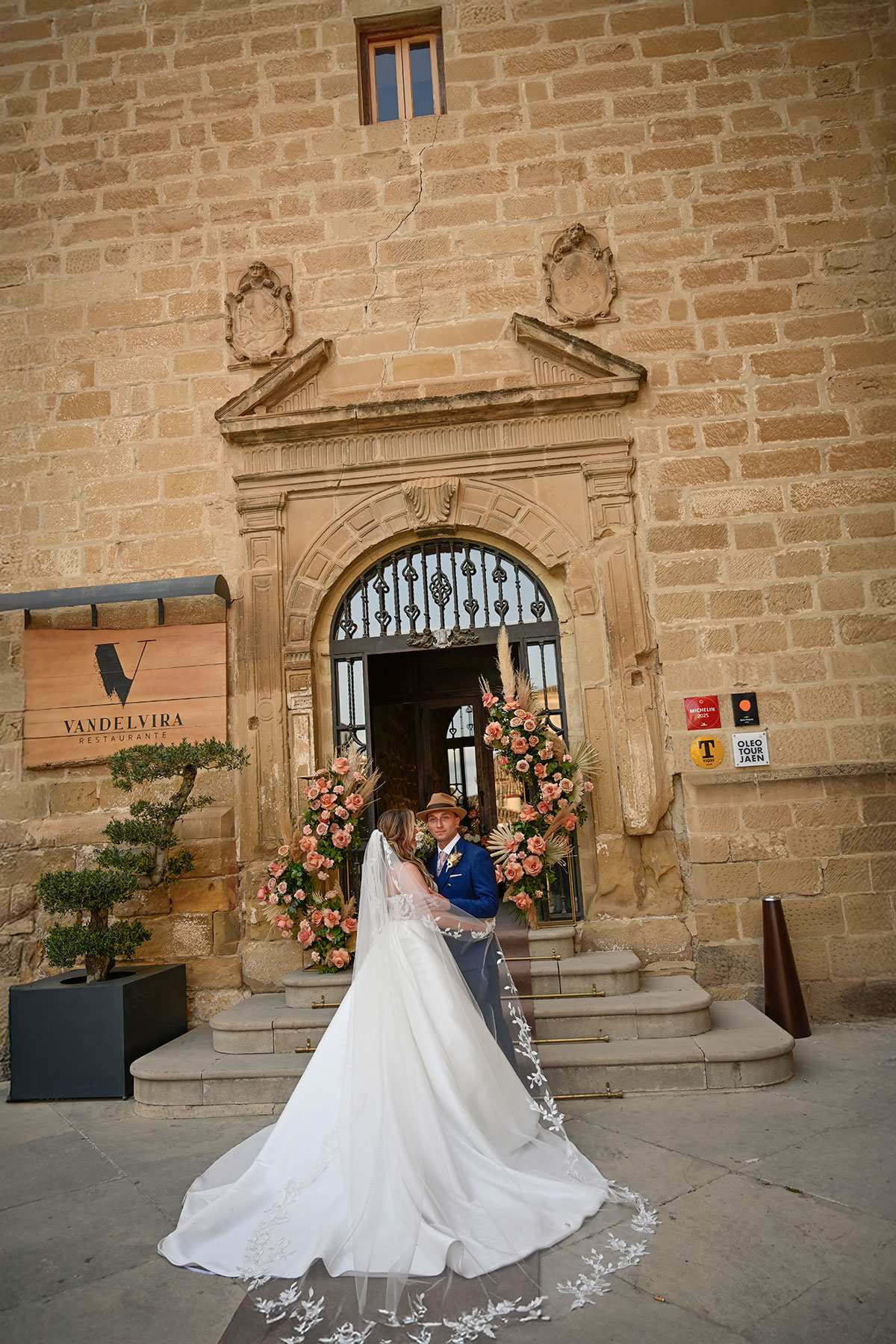 bride in trailing wedding dress with long veil stands with groom in fedora hat and blue suit outside brick building with pink flower displays at the entrance