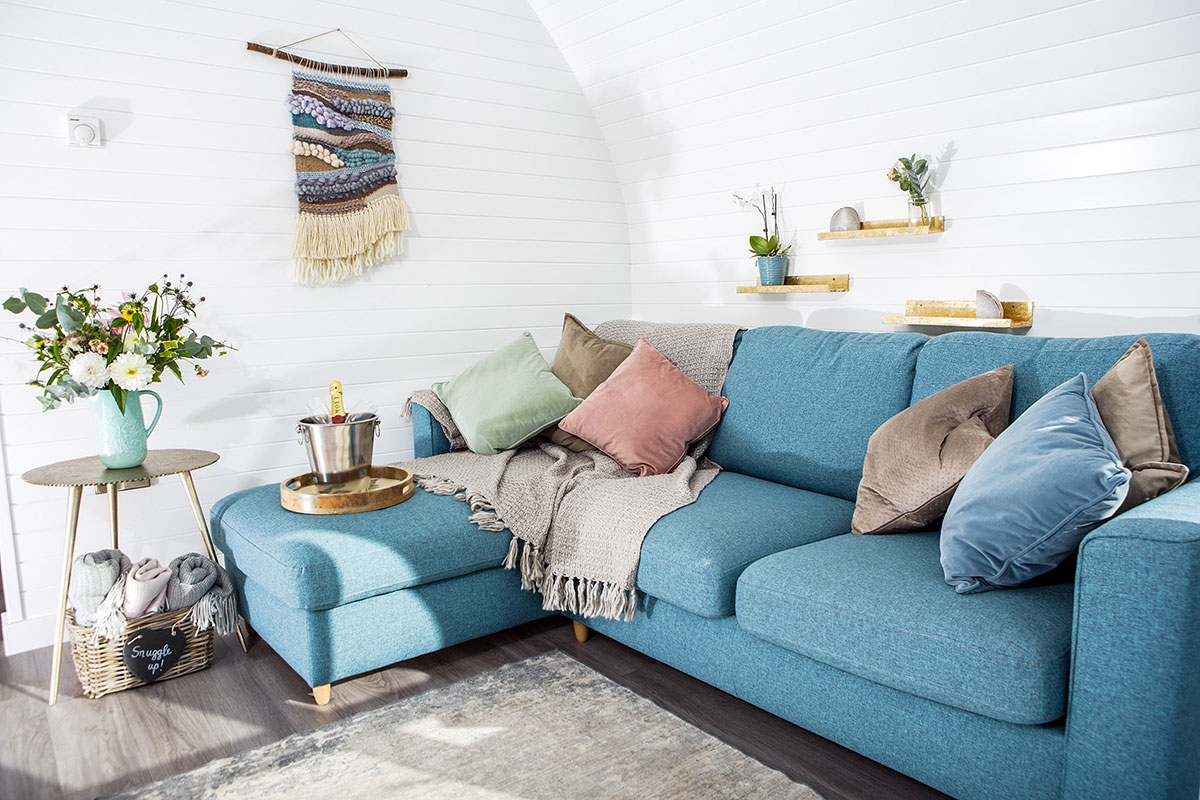 a blue L-shaped sofa covered in throw pillows and a tray with a bucket of champagne sits in the corner of a white panelled room with a side table with a jug of flowers
