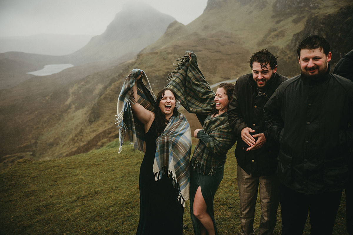 Wedding guests wrapped in tartan blankets laugh together on a windswept mountainside during a rainy outdoor celebration