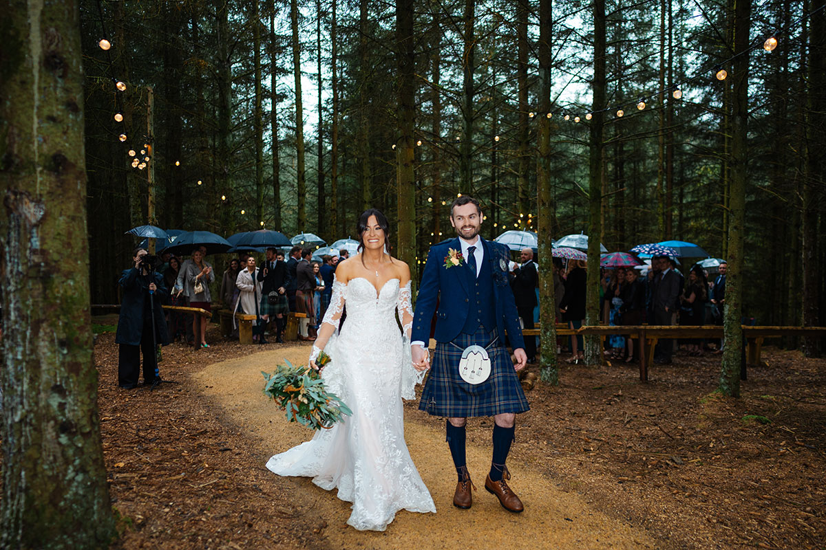 Newlywed couple walking through a woodland ceremony area at Eden Leisure Village outdoor wedding venue.