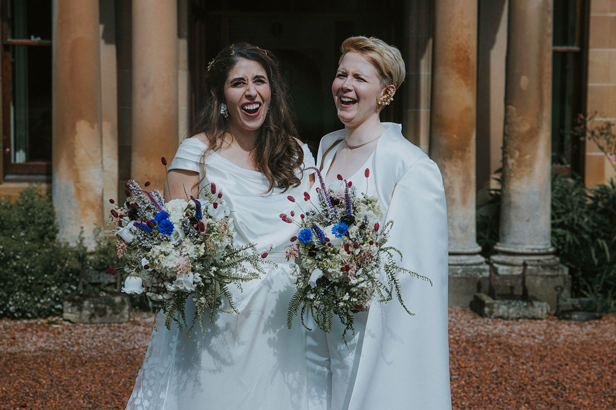 two brides laugh for a photograph on wedding day at cambo house while holding bouquets