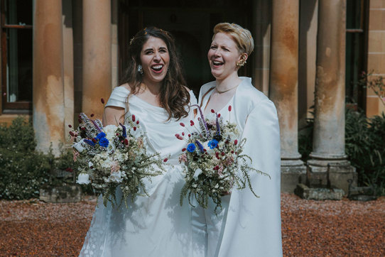 two brides laugh for a photograph on wedding day at cambo house while holding bouquets