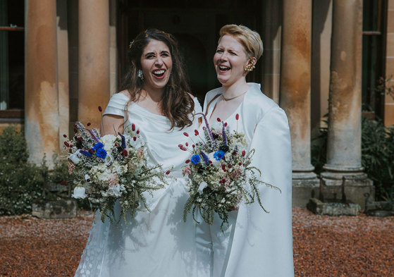 two brides laugh for a photograph on wedding day at cambo house while holding bouquets