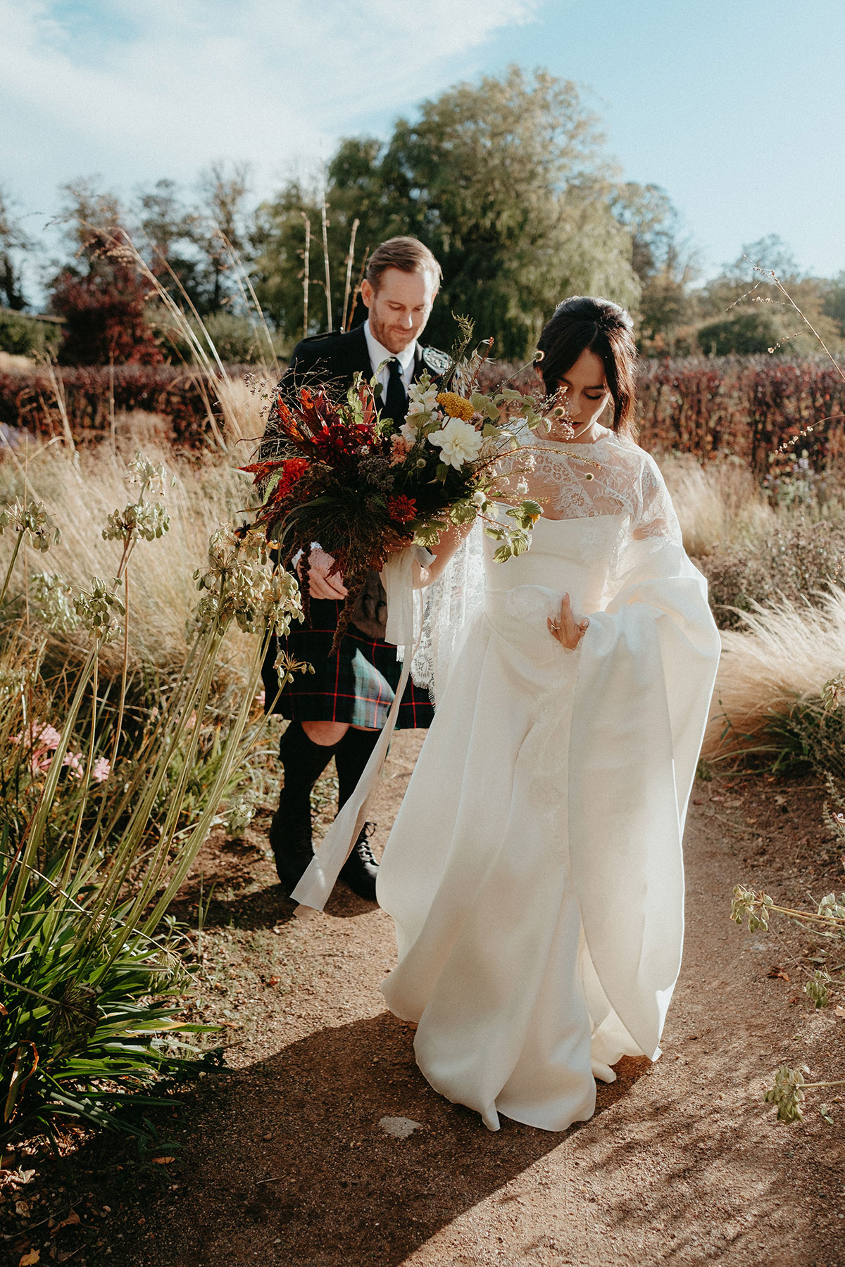 Bride and groom stroll through the Cambo Country House walled garden carrying colourful autumn bouquets on their wedding day in Fife.