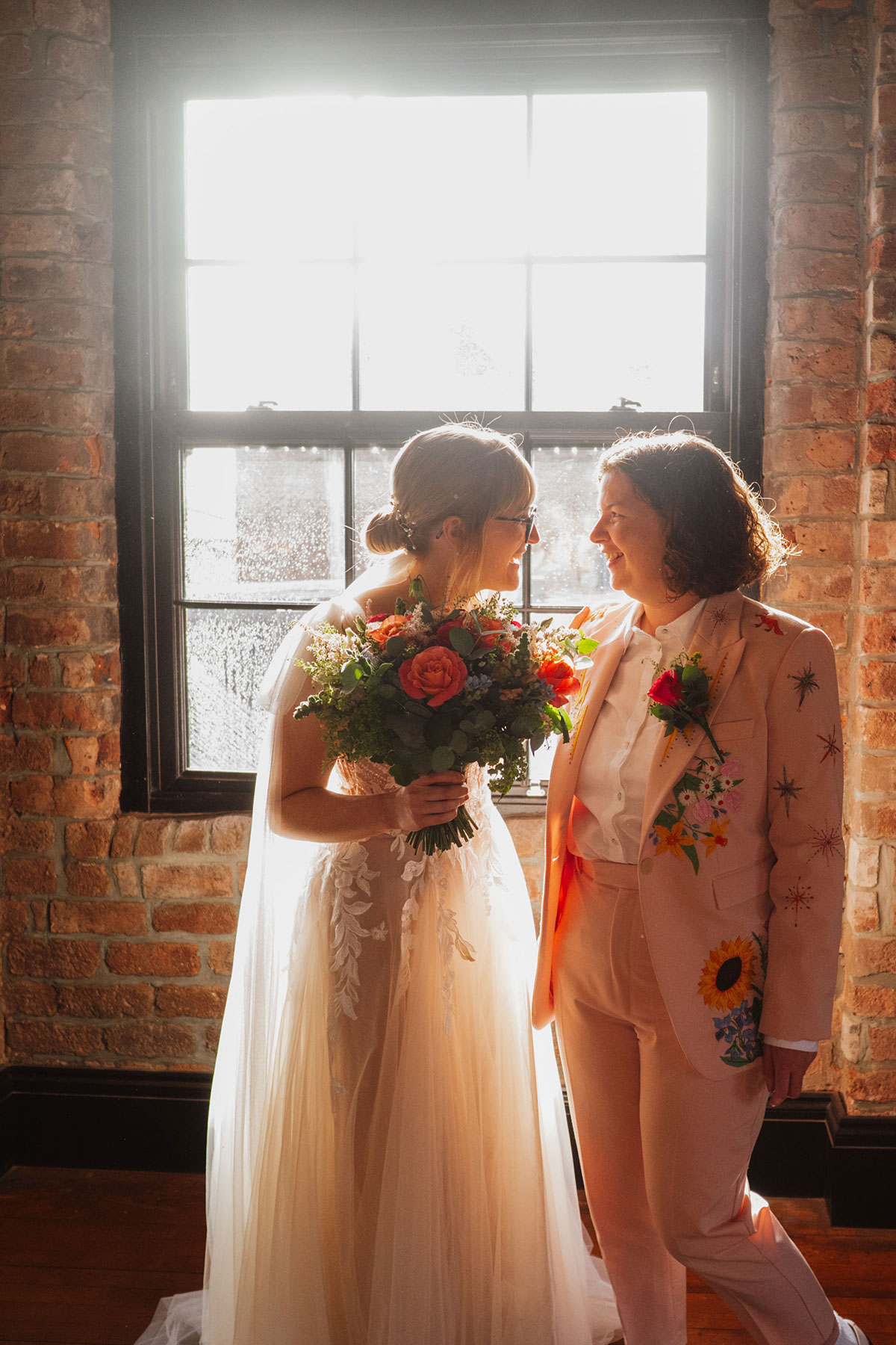 Brides standing close together in warm window light at the Engine Works, holding a vibrant bouquet.