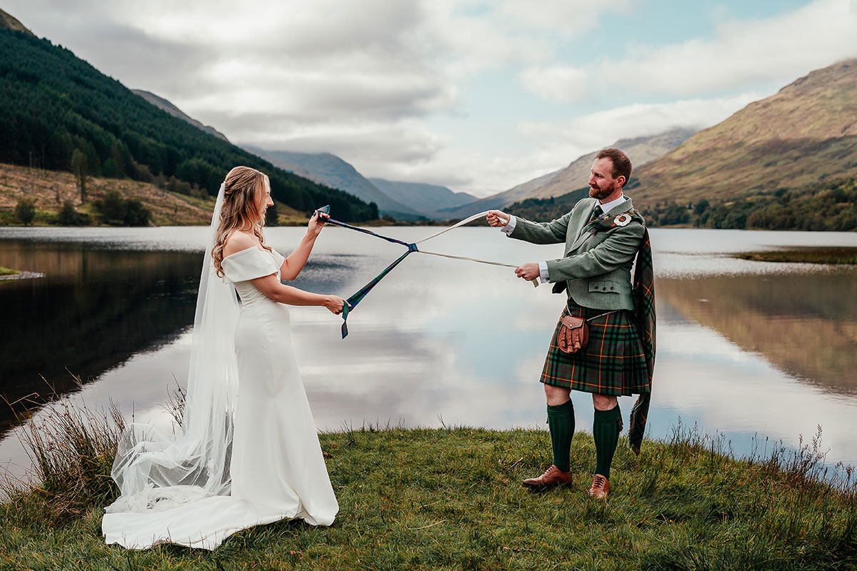 Bride and groom take part in a handfasting ceremony beside a tranquil loch with mountains reflected in the water