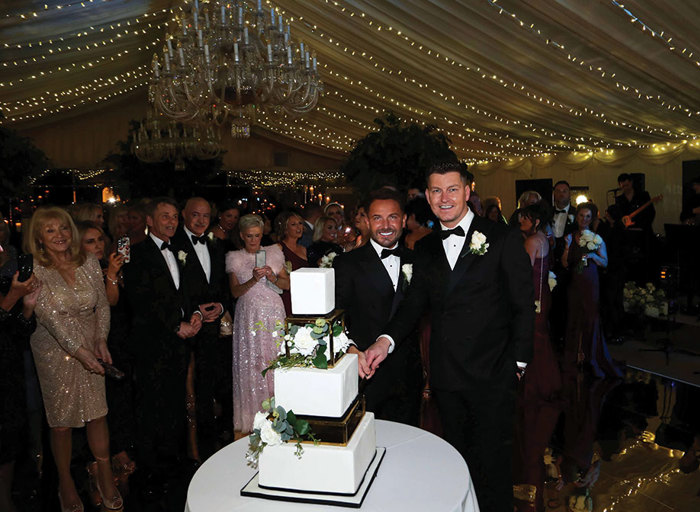 two grooms cutting a black and white square tier wedding cake below a canvas ceiling that's covered in fairy lights. Guests look on and take pictures