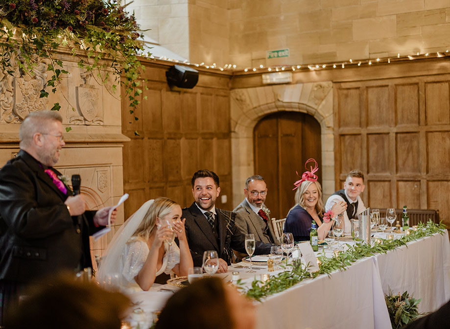 a top table with bride, groom and wedding guests watching on and laughing as person stands holding a microphone. There is a wooden panelled wall and stone fireplace in the background