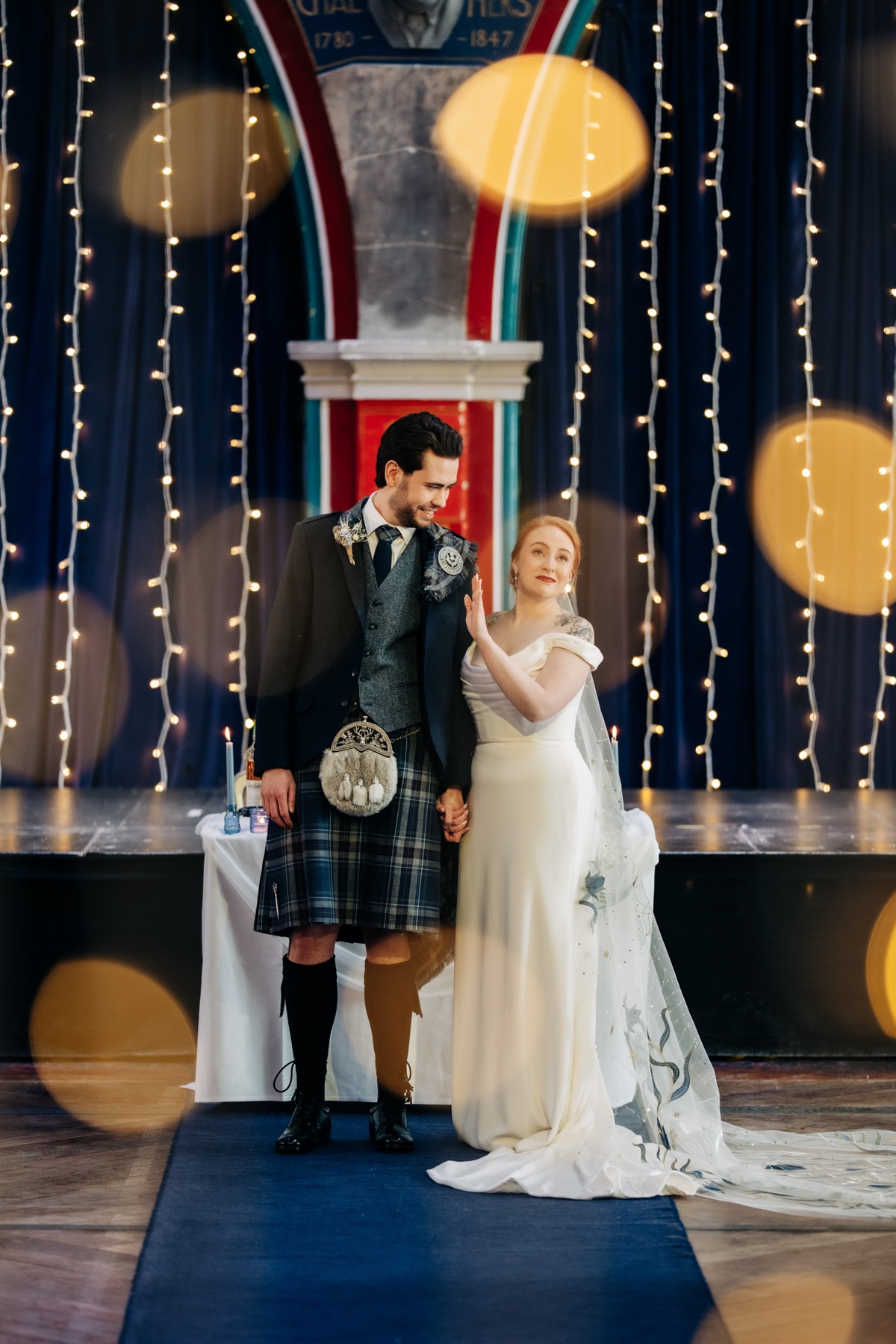 Robyn and James stand together during their wedding ceremony in Òran Mór’s Auditorium, with fairy lights and blue décor behind them.