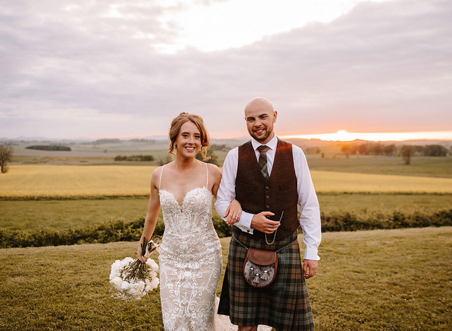 a bride and groom walking arm in arm through a grassy field at sunset
