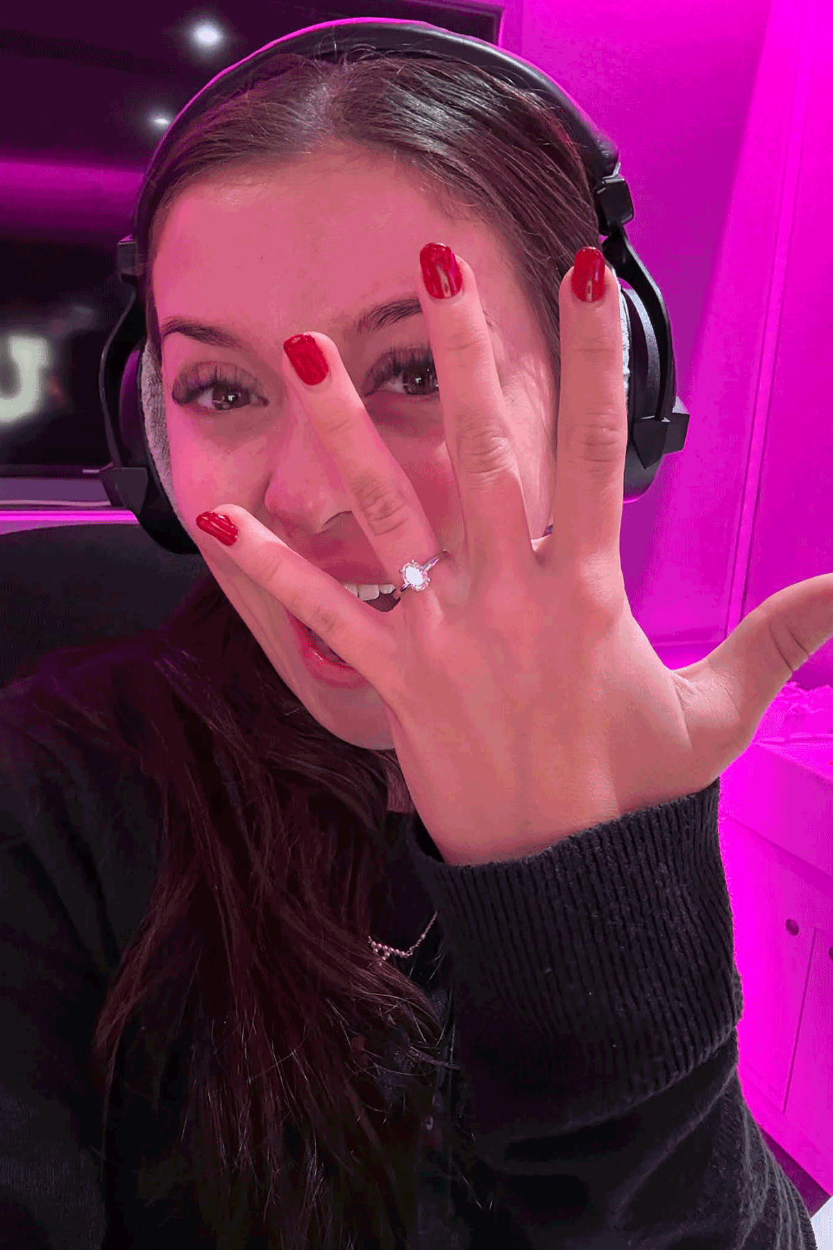 Bride-to-be wearing headphones and showing diamond engagement ring under pink studio lighting