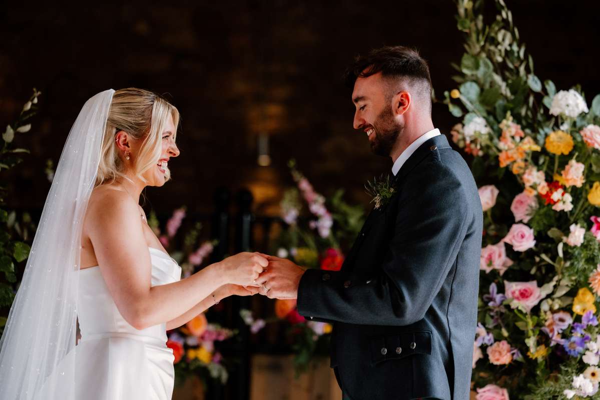 Bride and groom exchanging wedding rings beneath colourful floral installations at Falside Mill wedding venue
