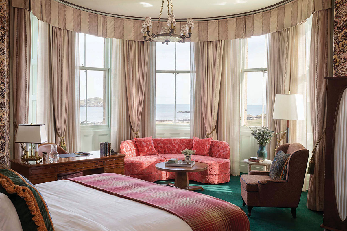 Bedroom with a large bay window overlooking the sea, featuring a bed, curved sofa, armchair and soft neutral furnishings