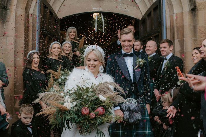 A bride and groom leave the church with their wedding party behind them as confetti is thrown over them