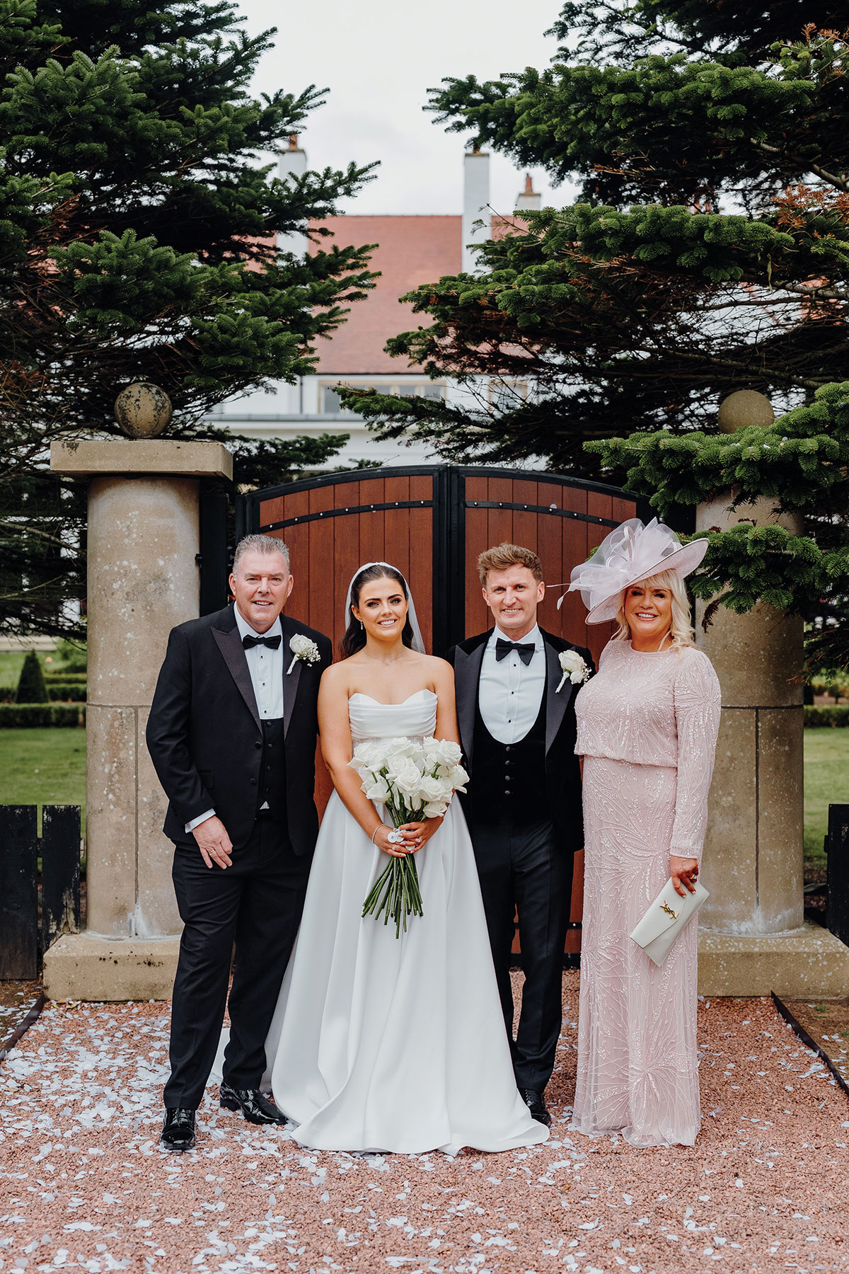 Bride Sophie poses outdoors with her parents and groom in front of wooden gates and evergreen trees, all dressed in formal wedding attire