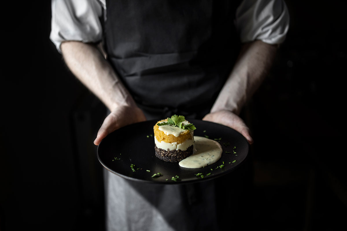 Close-up of a chef holding a black plate with a layered dish topped with herbs and served with a creamy sauce