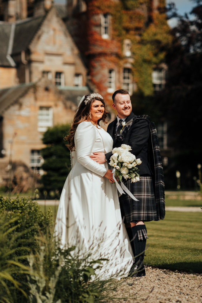 A man in a dark kilt and a bride in a long sleeved wedding dress stand with their arms around each other outside