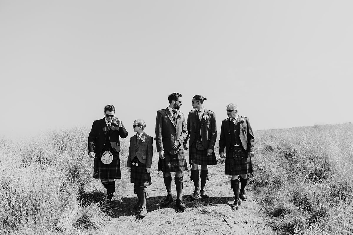 black and white picture of groom and groomsmen walking down sandy hill towards the beach