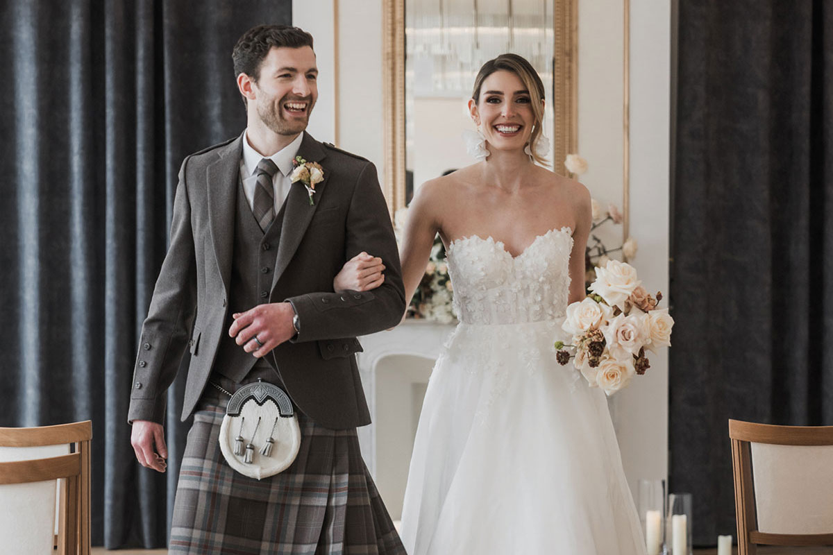 a bride and groom walking arm in arm up the aisle at the Exchange wedding venue in Glasgow. The brunette bride is wearing a wedding dress with floral applique on the bodice and the groom is wearing a brown tartan kilt outfit.