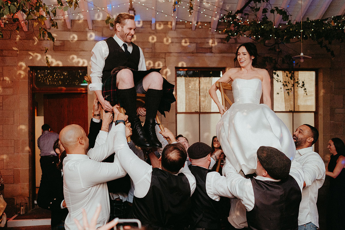 Guests lift the bride and groom on chairs during the lively Jewish hora dance at the Cambo Country House wedding reception.