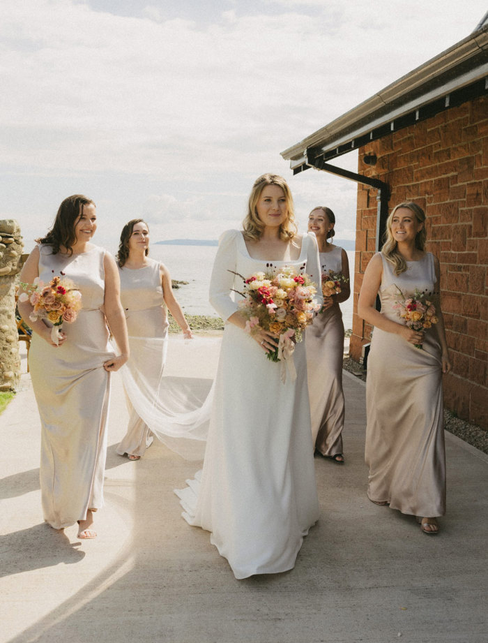 a smiling bride walking with four bridesmaids outside a red brick building in the sunshine