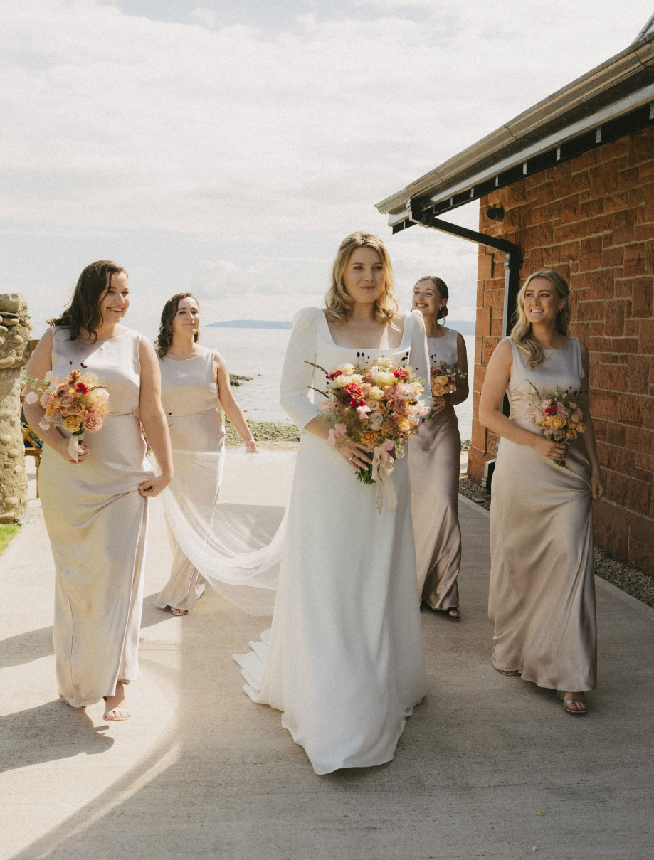 a smiling bride walking with four bridesmaids outside a red brick building in the sunshine