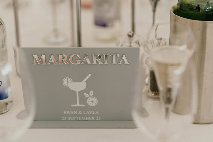 A grey sign with the word 'Margarita' in silver at the top and a picture of a cocktail and citrus fruit underneath in white. Also in white below that is the text 'Ewan & Layla 23 September 23'.
