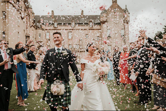 A bride and groom walk towards the camera with a castle behind them as their guests throw pink and white confetti over them 