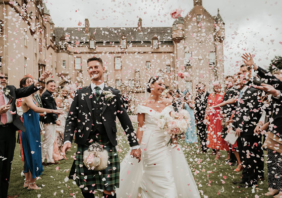 A bride and groom walk towards the camera with a castle behind them as their guests throw pink and white confetti over them 